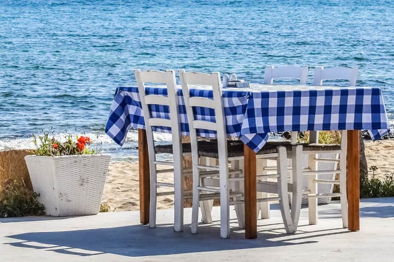 Une table avec un tissu à carreaux bleu-blanc et deux chaises blanches au bord de la mer.