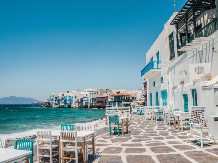 the cobbled promenade of Little Venice in Mykonos with the colourful buildings above the water and some restaurant tables next to the shore