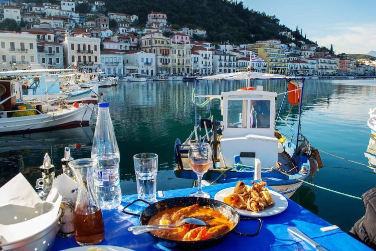 a table next to a coastline in Peloponnese, Greece, with a plate of shrimps with red sauce and feta cheese, a plate of fried squid, red wine and water